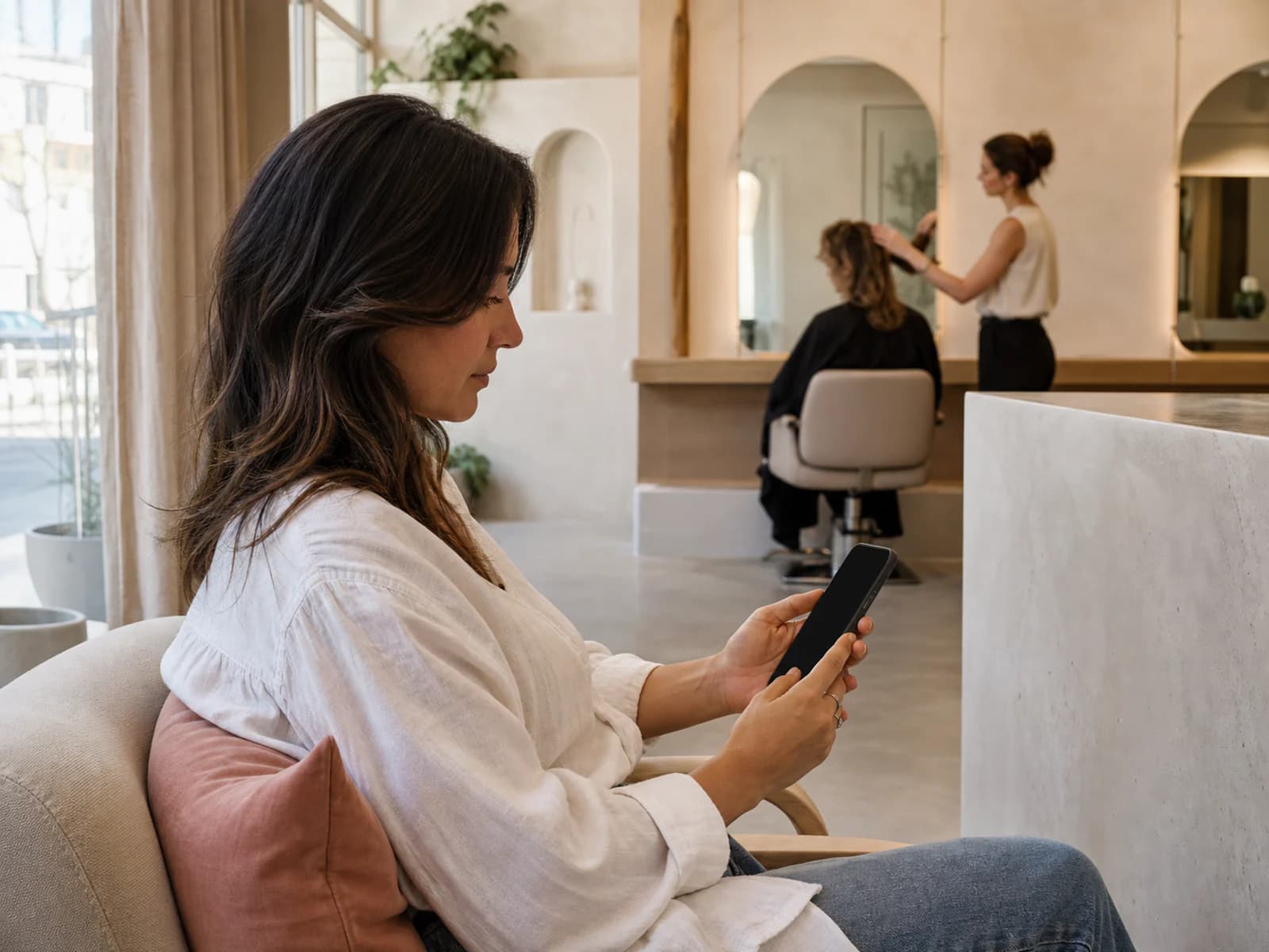 Salon customer browsing a phone with a blank screen while a stylist works in the background